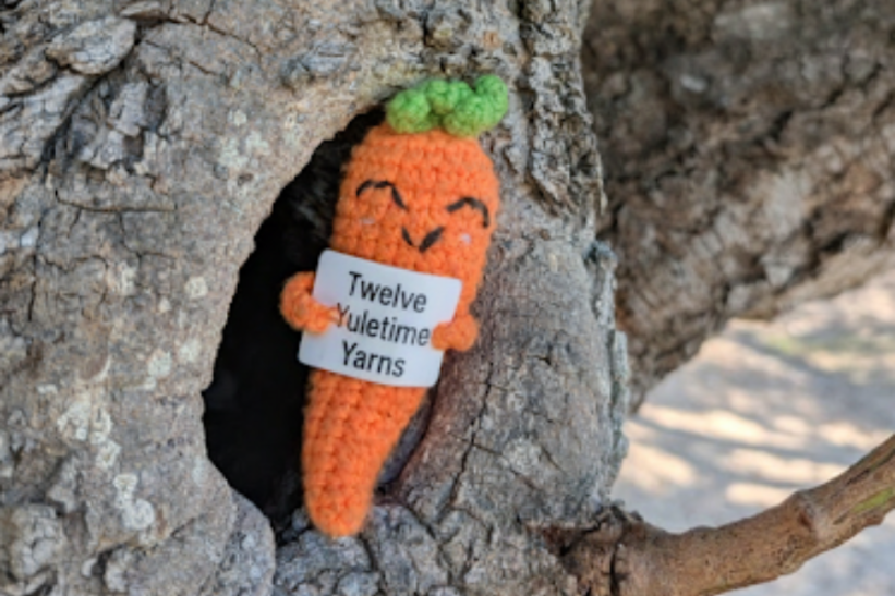 A crocheted carrot in a tree, holding a sign that reads 'Twelve Yuletide Yarns'